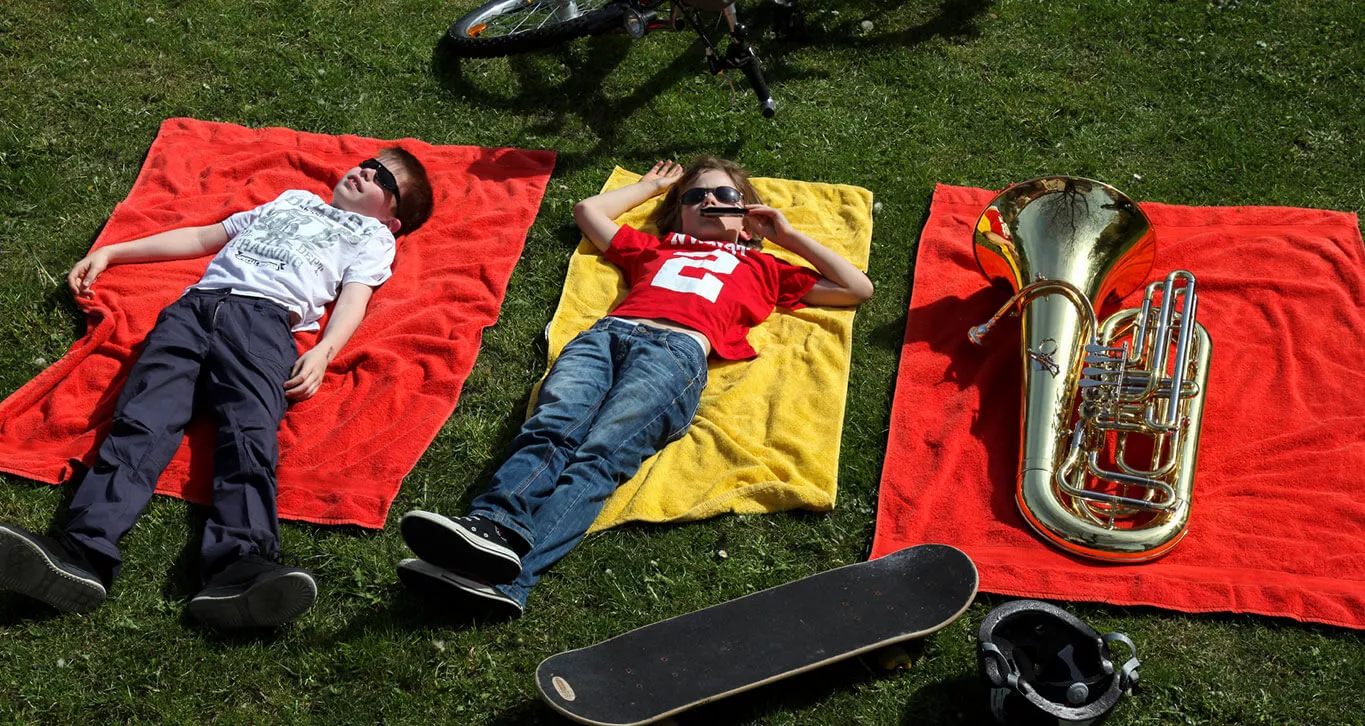 Zwei Jungen liegen mit einer Tuba auf Handtüchern auf einer Wiese.