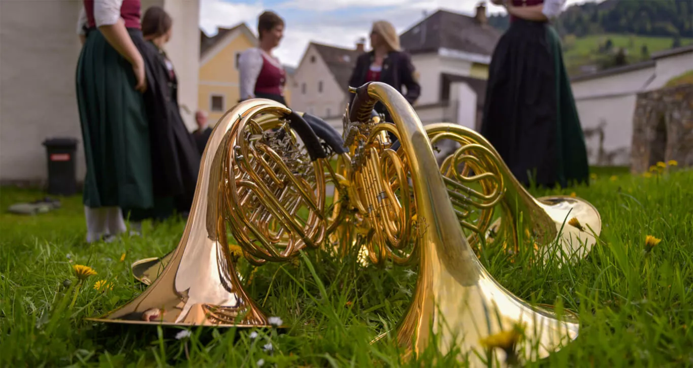 Im Vordergrund liegen drei Walhörner auf einer Wiese, im Hintergrund stehen Frauen in Dirndl.